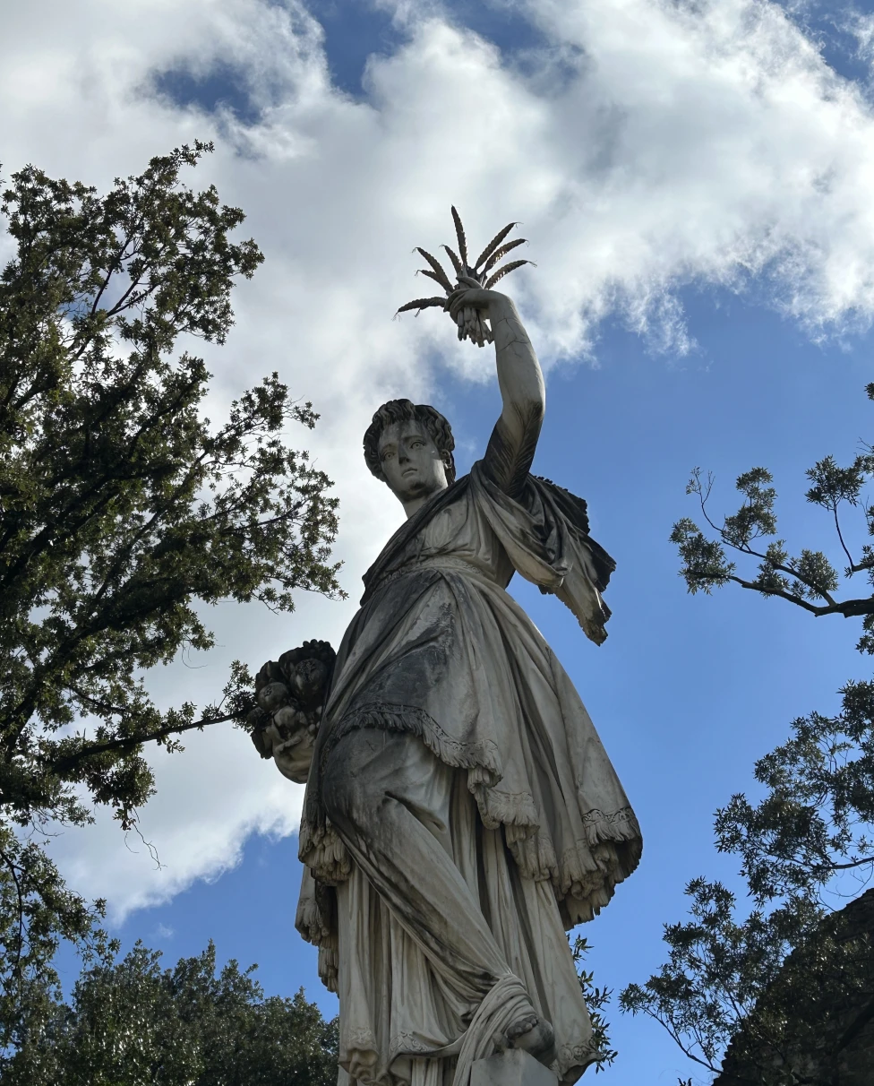 An ornate tatue with a cross, bathed in sunlight, towering over trees under clear blue skies.