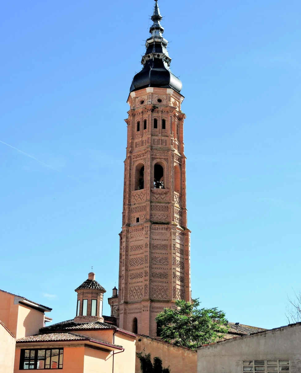 A tall ornate tower with a spire, featuring intricate brickwork patterns, set against a clear blue sky.