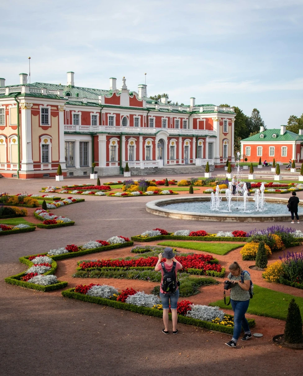 Kadriorg Park is the most outstanding palatial and urban park in Estonia. Picture features manicured gardens and a circular fountain in front of a large red building with white trim and green roofs.