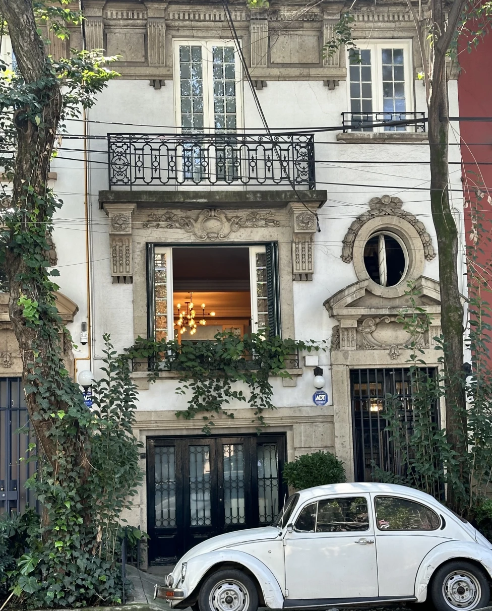A vintage white Volkswagen Beetle parked in front of an old building with greenery and a lit interior.