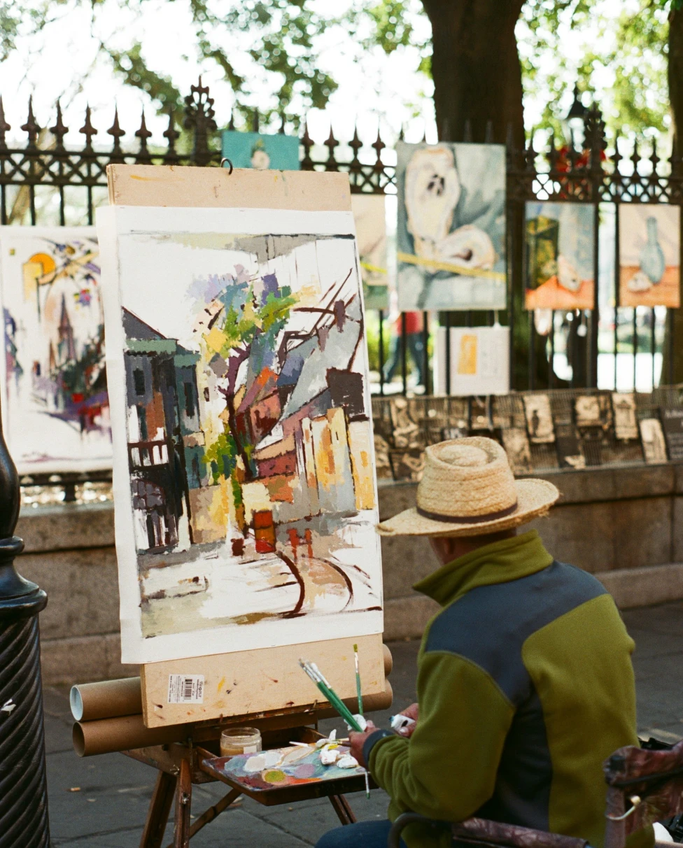 A man wearing a straw hat and green jacket, sitting on a stool and painting a picture outside.