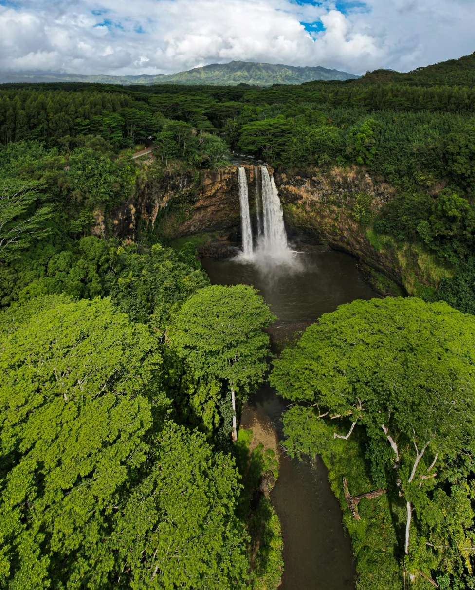 A waterfall, lush greenery and mountains under a partly cloudy sky.
