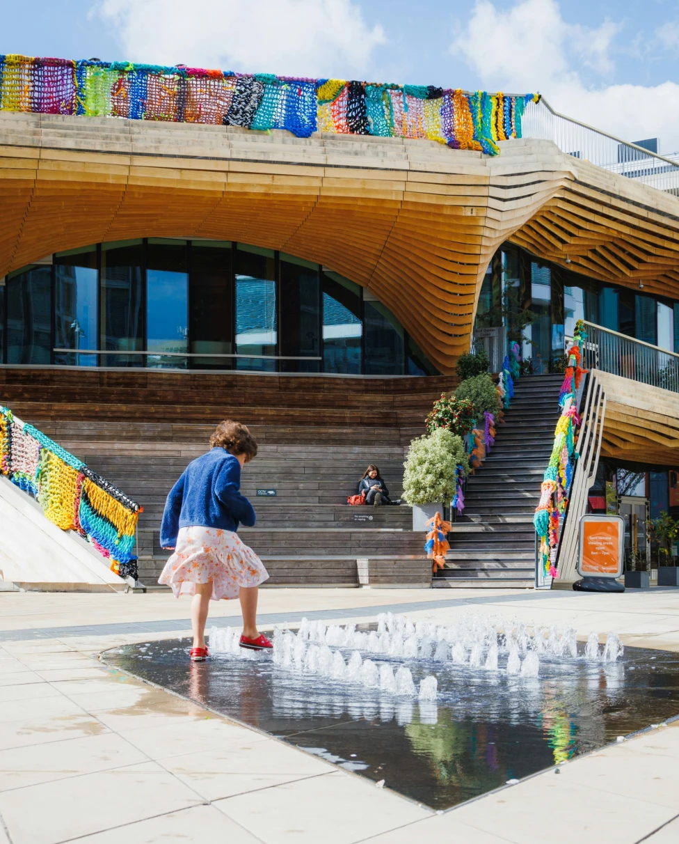 A modern building with a wooden facade and colorful banners, featuring a fountain and people sitting on steps.
