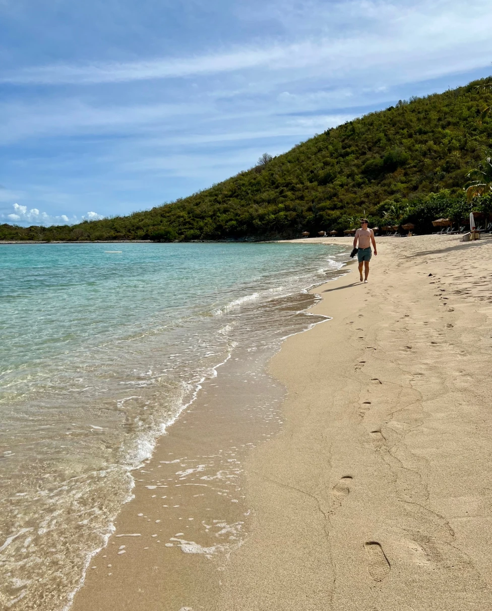 A man walking down a beach with crystal clear water and a green mountain in the background.