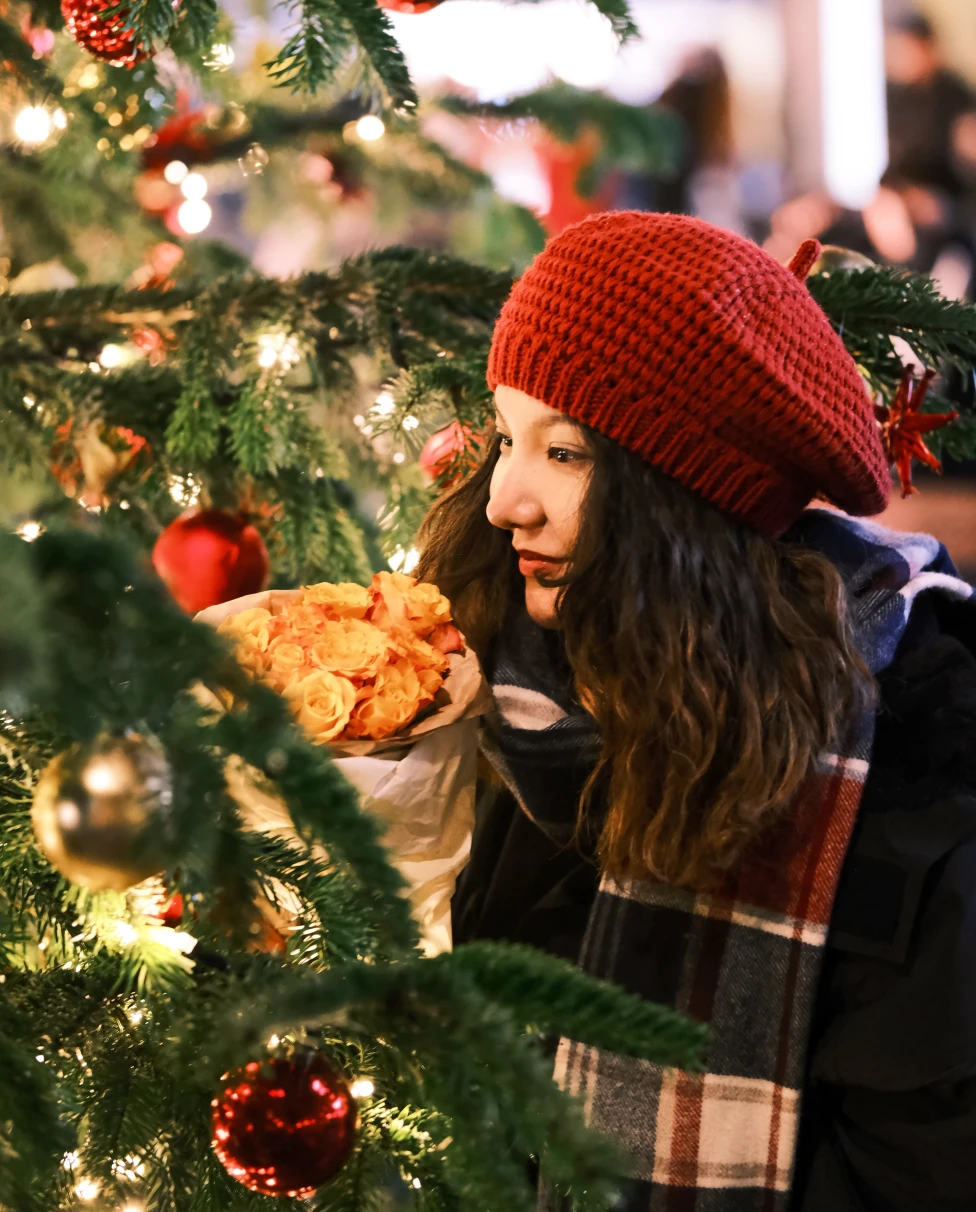 A girl in a red hat standing next to a Christmas tree at a market.