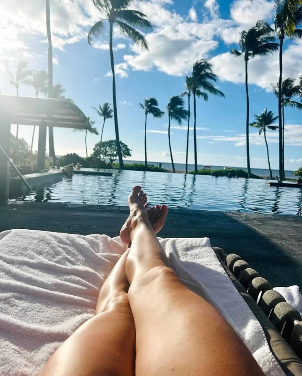 A pool with palm trees and legs of a person taking the picture lounging on pool chair.