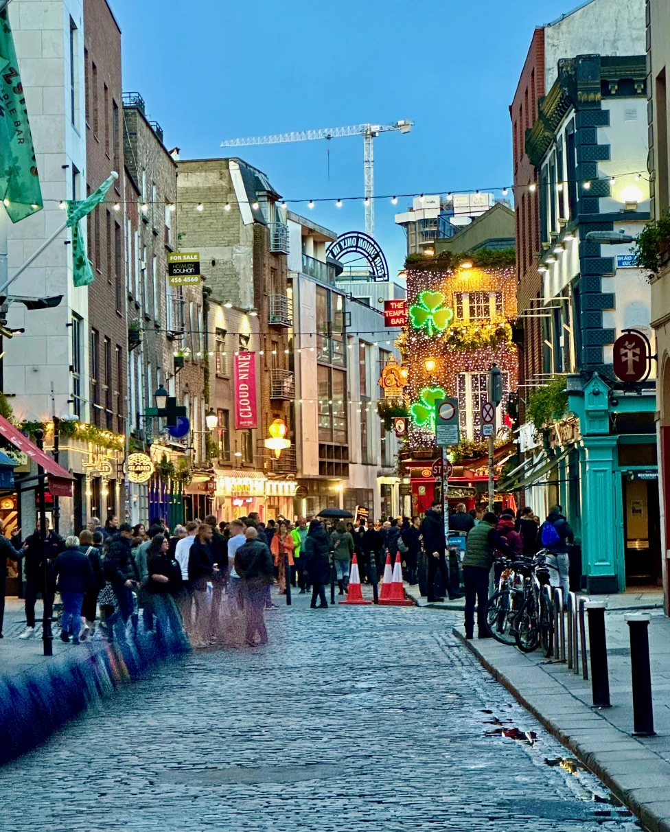 Cobblestone city streets in Dublin in the evening with neon shamrocks and people walking around.