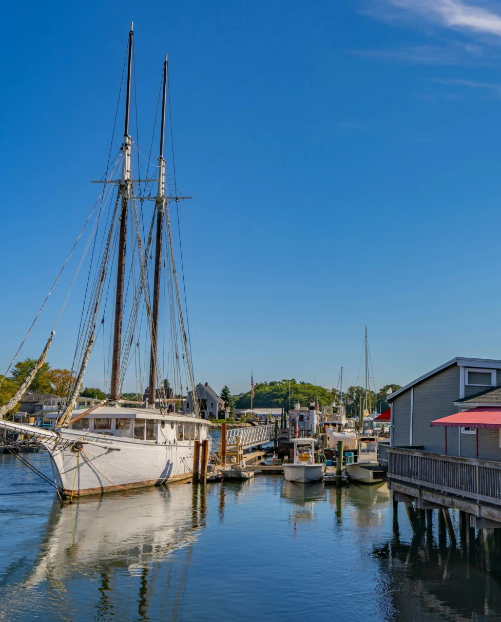 The image depicts a scenic harbor with a tall ship and “The Black Shack” waterfront building under a clear blue sky.