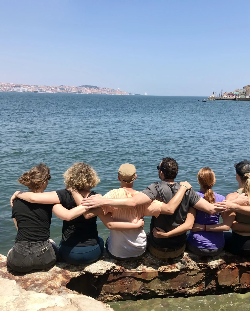 A group of people sitting on the waters edge with arms around each other, backs to the camera