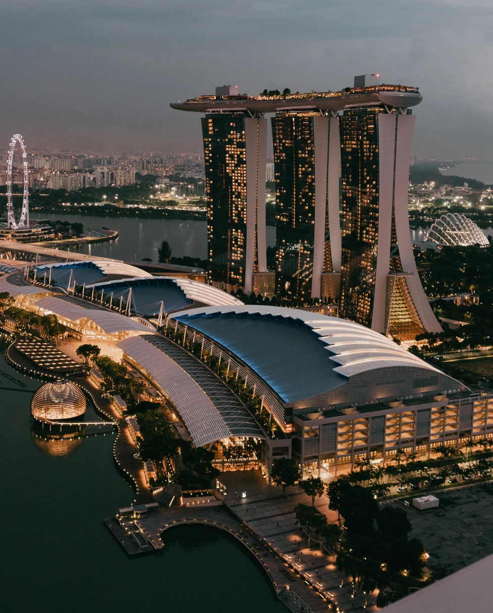 three tall buildings lit up at night on the water surrounded by sprawling modern city