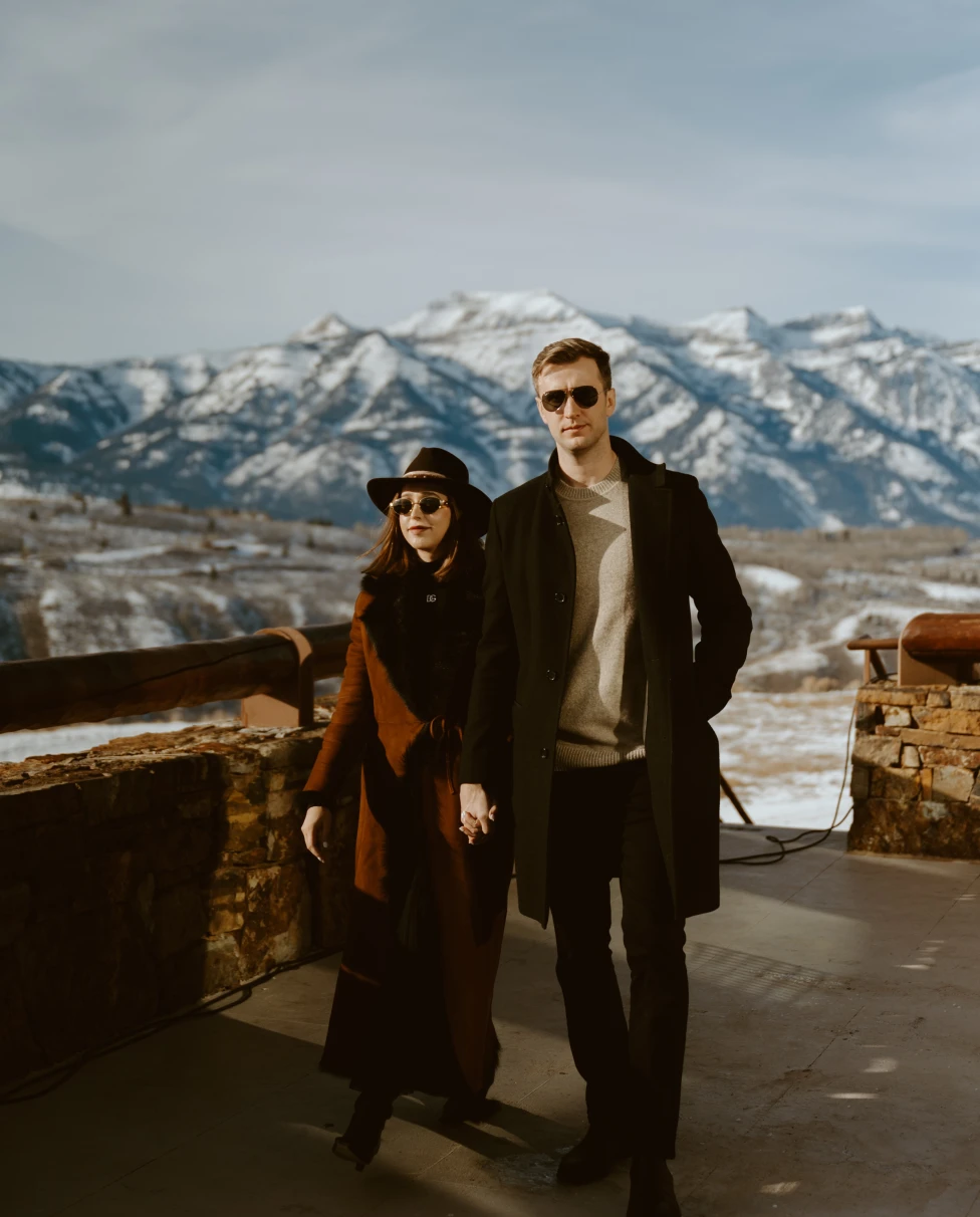 A couple dressed in swanky winter clothes, standing on an open balcony, posing in front of snow-covered mountains.