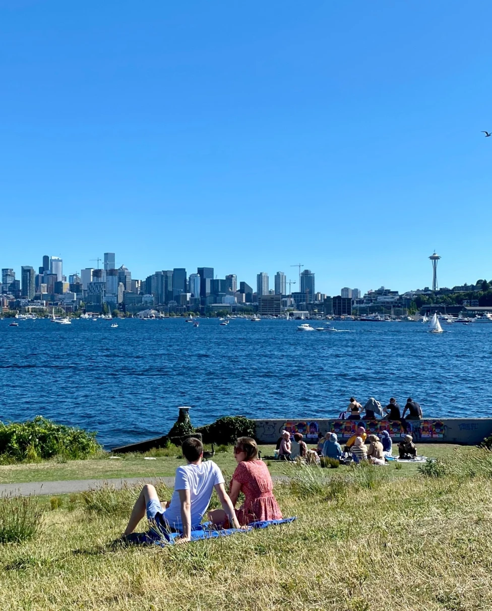 A picture of people sitting on a grassy lawn with a view of the sea and Seattle skyline in the distance.
