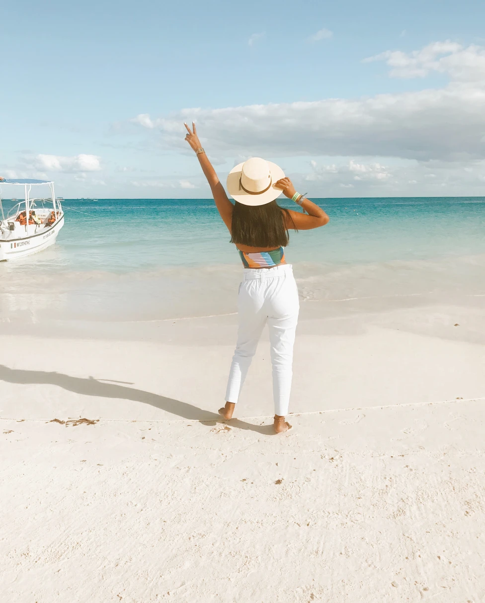 Girl with white brimmed hat stands with arm in the air by the ocean in Tulum, Mexico