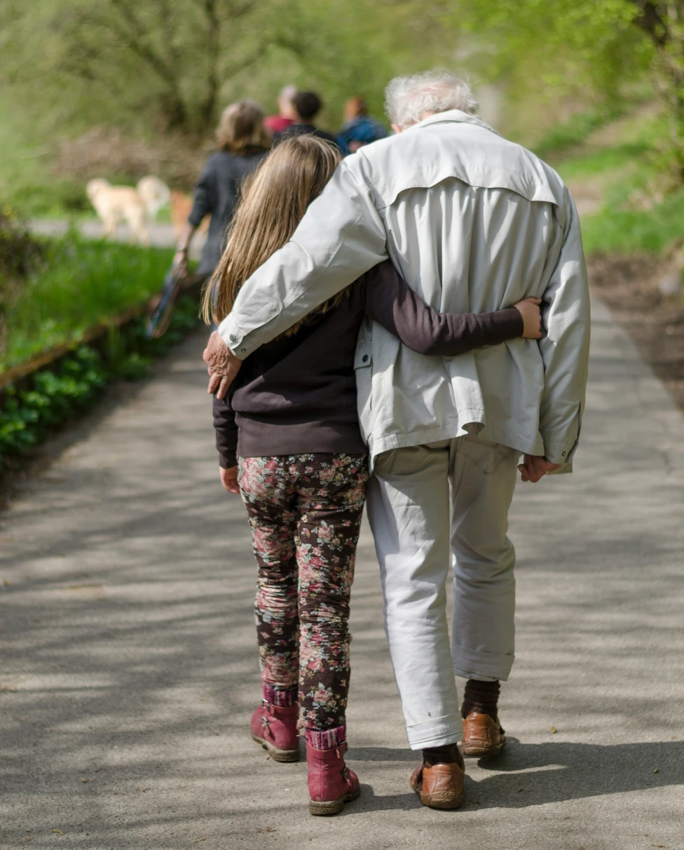 Two companions walk arm in arm along a path surrounded by lush greenery.