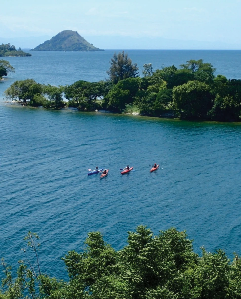 Kayakers on the beautiful Lake Kivu, a large blue water body with strips of trees jutting into it and a mountain in the distance.