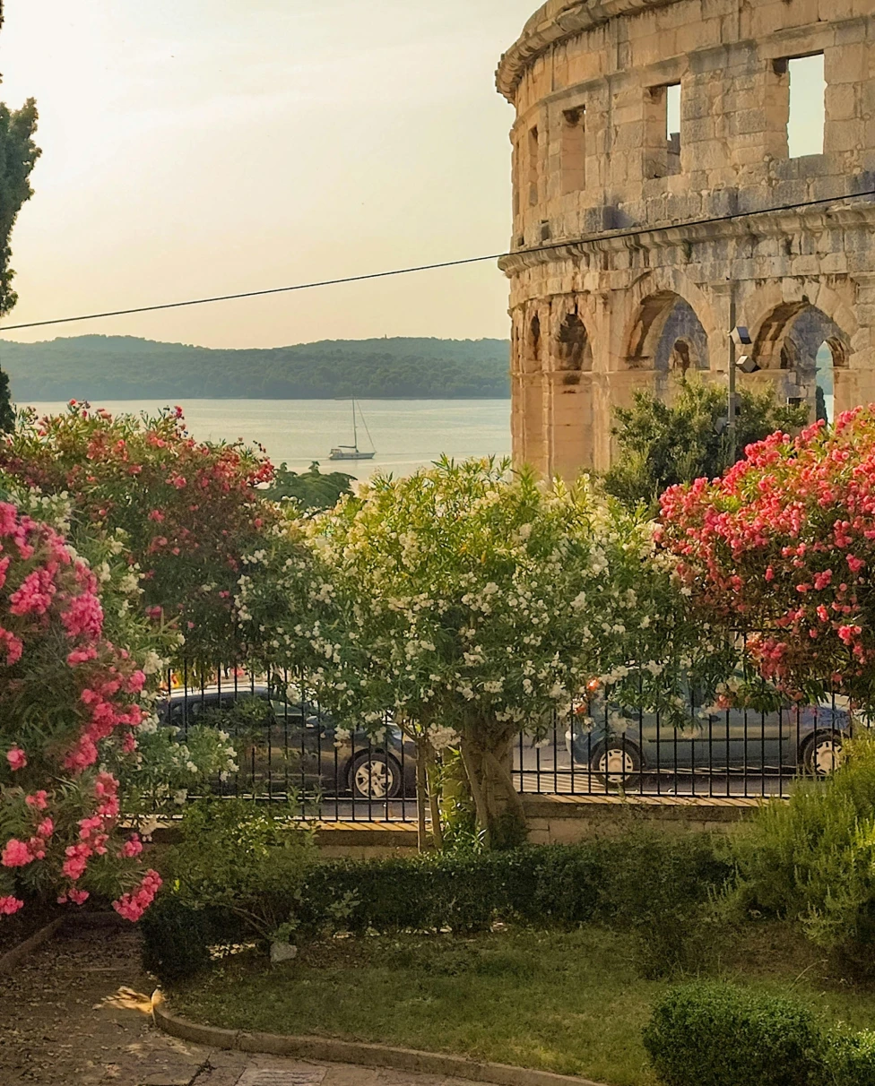 A side view of the exterior of the Roman amphitheater in Pula, with flowers and bushes in the forefront, and sea and mountains in the background.