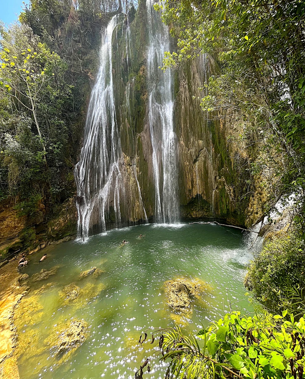 A stunning waterfall cascades into a clear green pool, surrounded by lush greenery on a bright sunny sky.