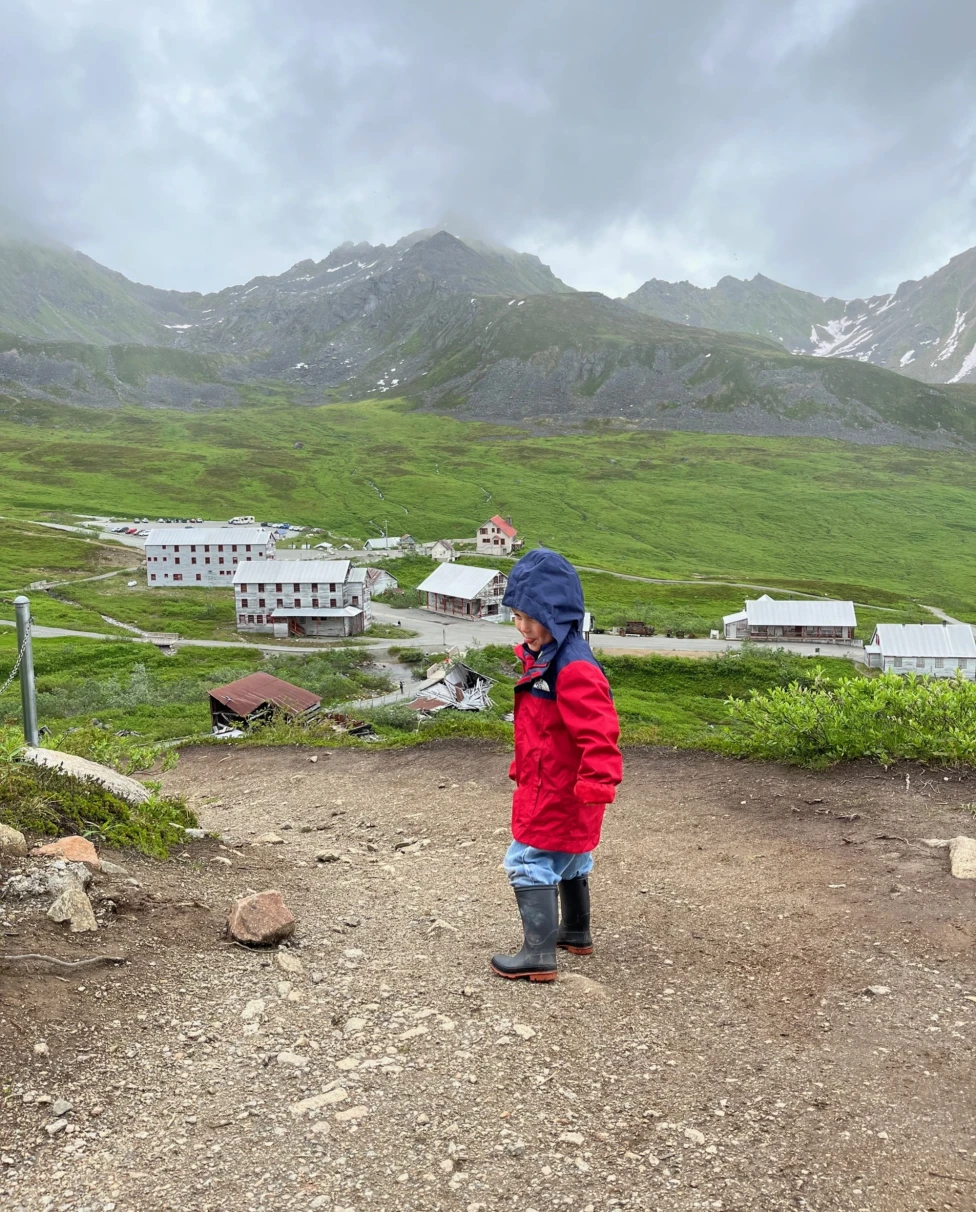 A child in red clothing stands on a mountainous terrain overlooking a valley with buildings, under a cloudy sky.