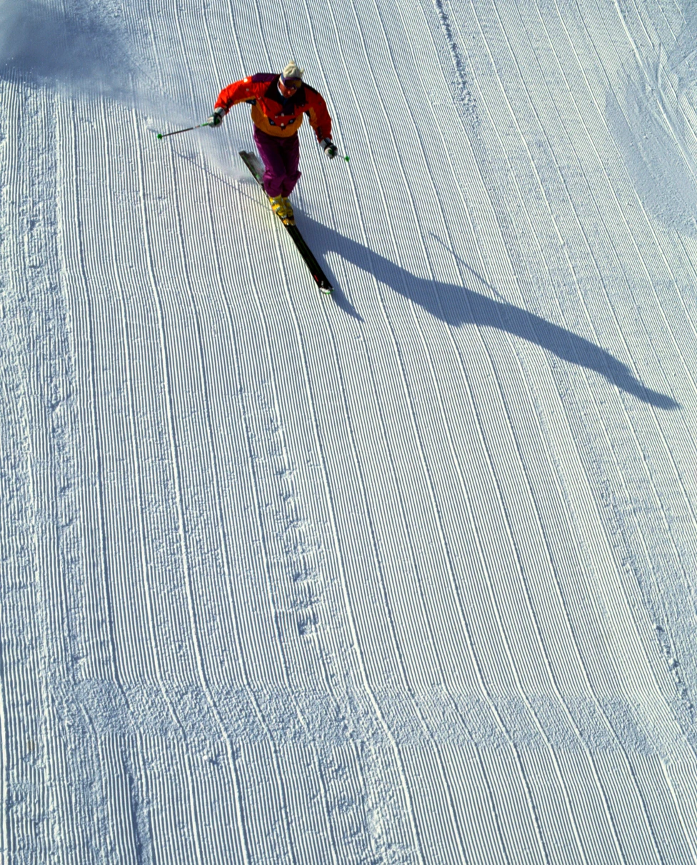 A skier in a red jacket ski's down a steep snowy slope on a bright day.