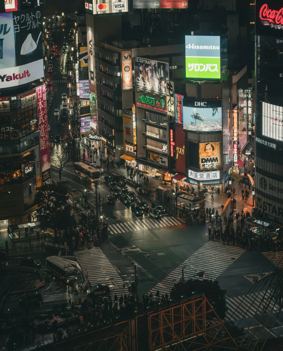 An image of an overview of Tokyo's city center with various lights, billboards and LED screens. There are people walking along the street down below.