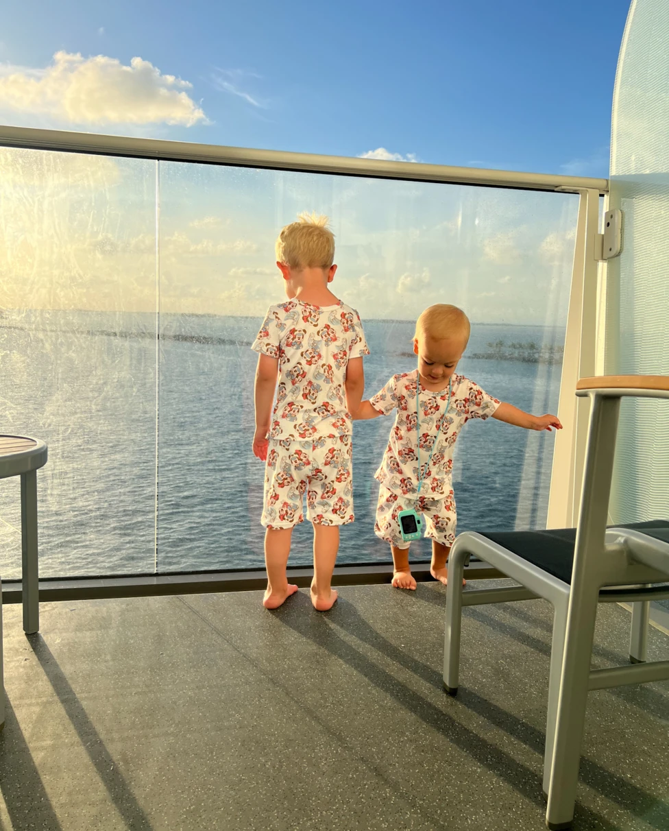 The image shows two kids in matching outfits, enjoying a sunny day by the sea behind a glass barrier on a cruise.
