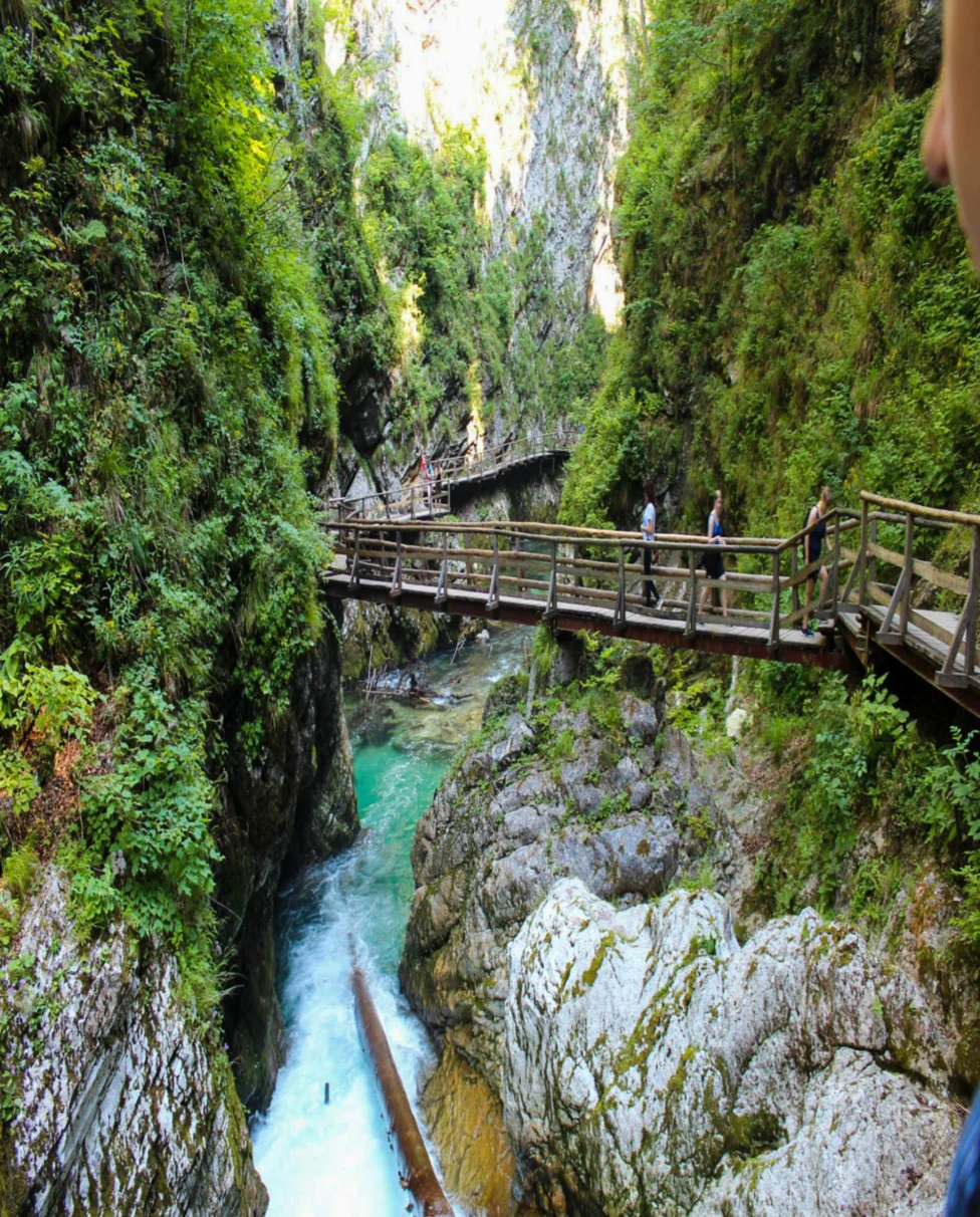 The image features a wooden walkway along a rocky gorge with a turquoise river below, amidst lush greenery and visitors enjoying the view.