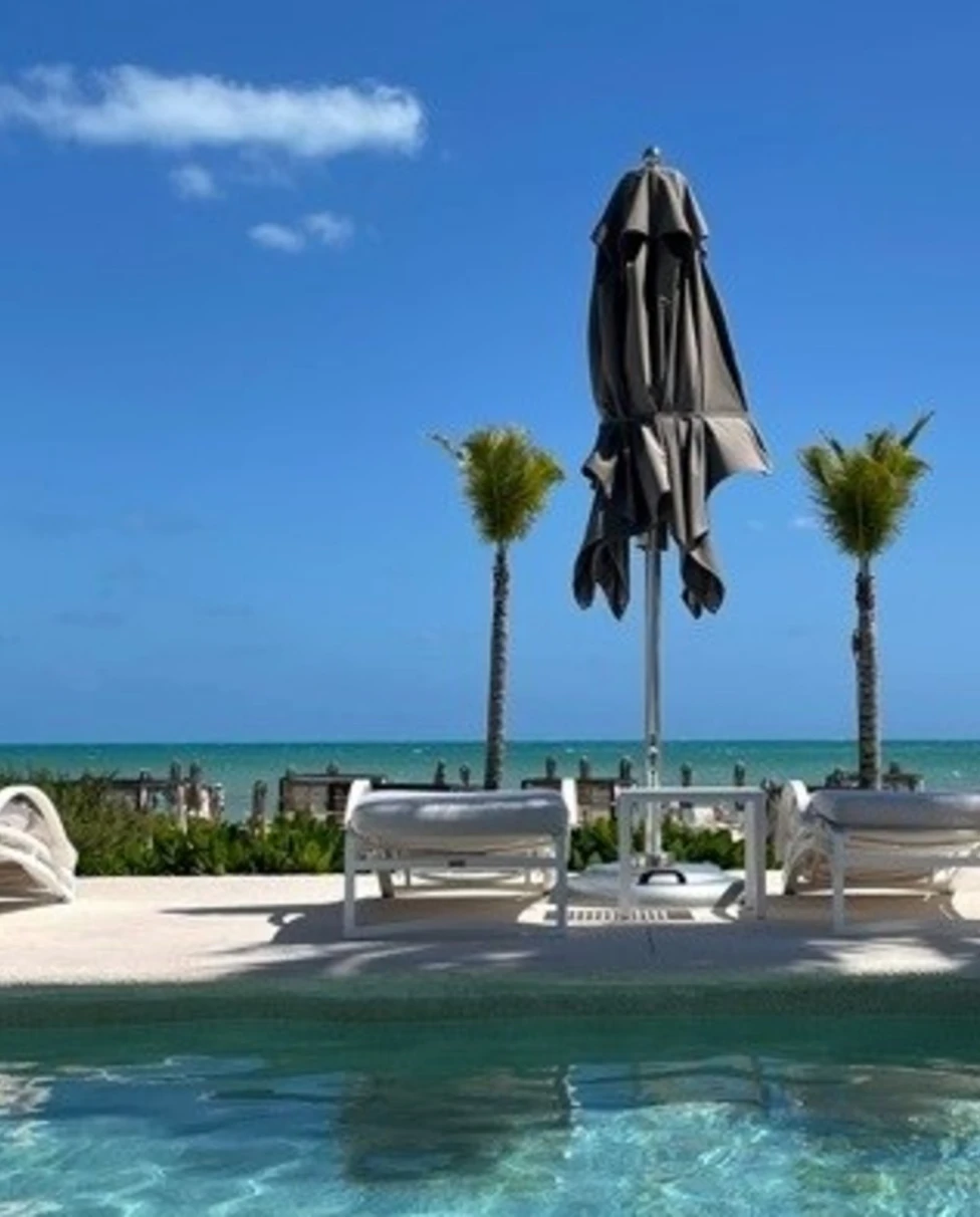 A tranquil poolside view with sun loungers, a closed umbrella and palm trees against a backdrop of a clear blue sky and ocean.