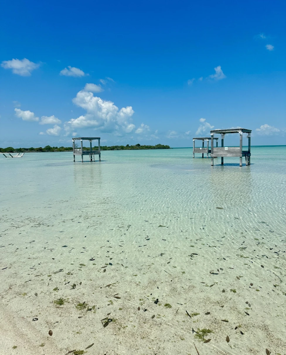 A clear shallow body of water with two dilapidated wooden structures under a blue sky.