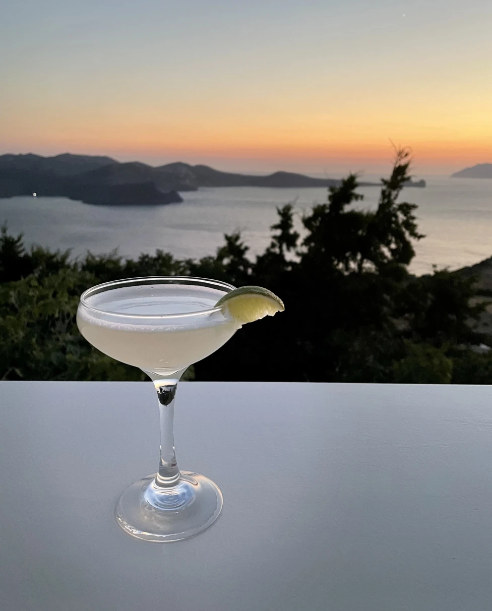A close-up of an elegant cocktail set on a table, with a view of the sunset over the sea in the background.