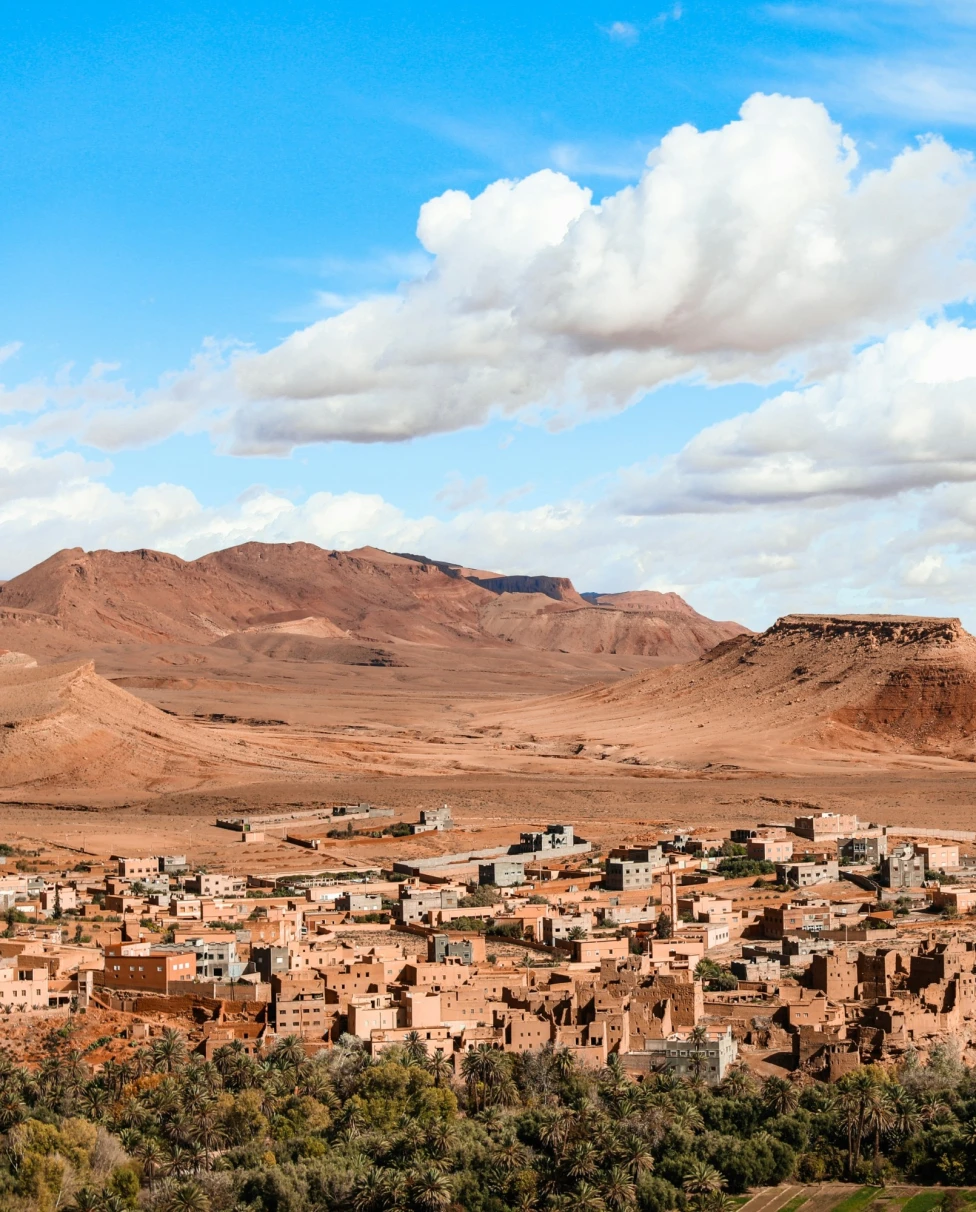 A traditional Moroccan village with clay buildings nestled at the foot of a hill under a blue sky with scattered clouds.