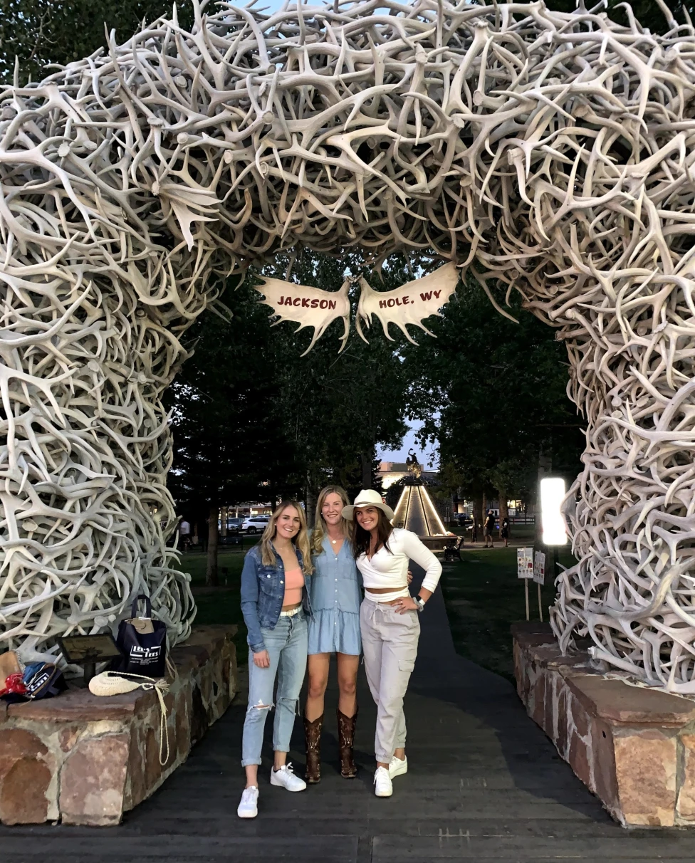 Three women standing below an outdoor archway made of antlers that says "Jackson Hole, WY" with trees in the background.