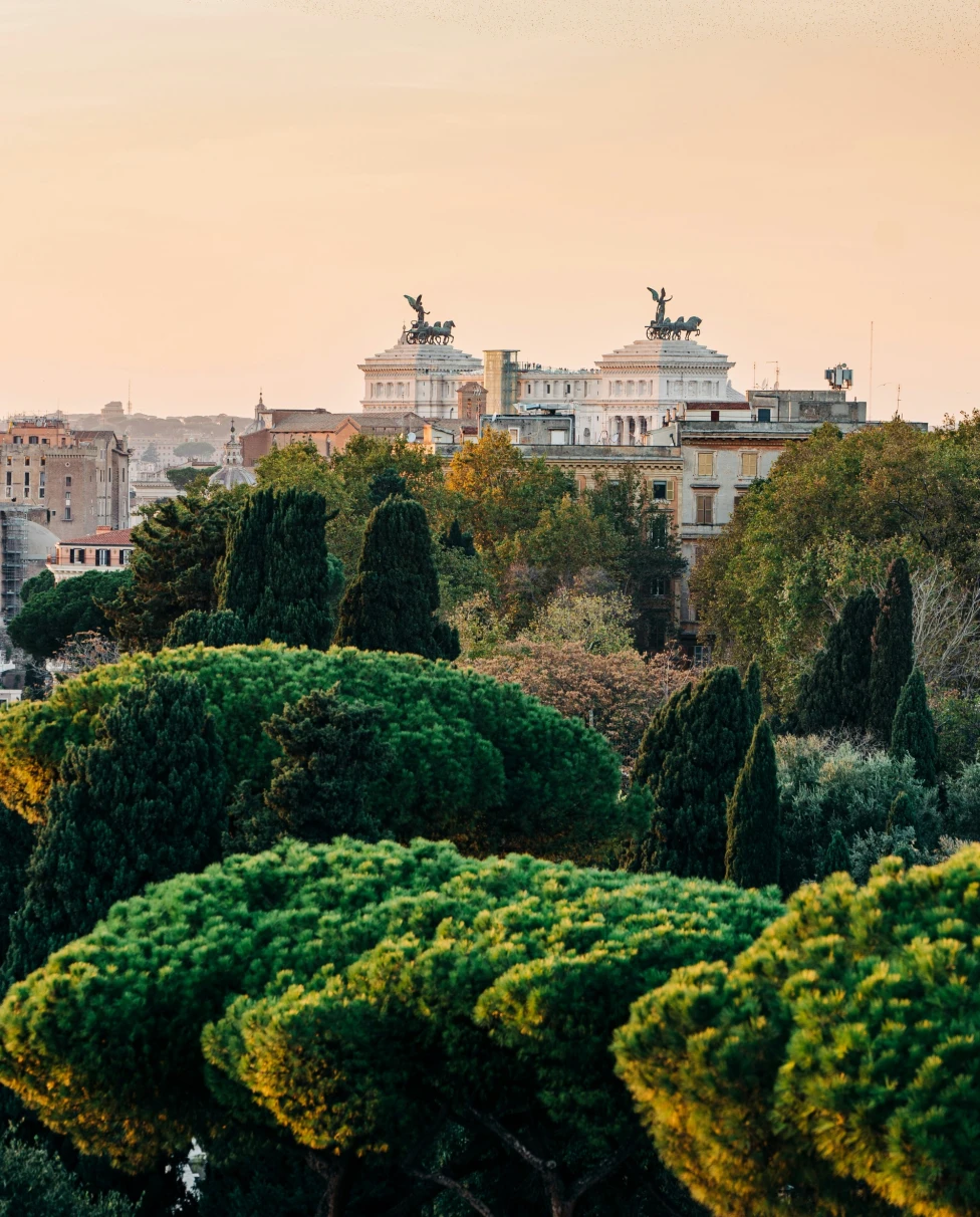 The image shows a beautiful cityscape at dusk, with buildings and statues silhouetted against a pastel sky and lush greenery in the foreground.