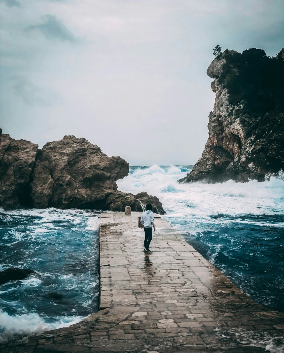 A picture of a woman in a white shirt and black pants on a concrete pathway near the sea during the daytime.