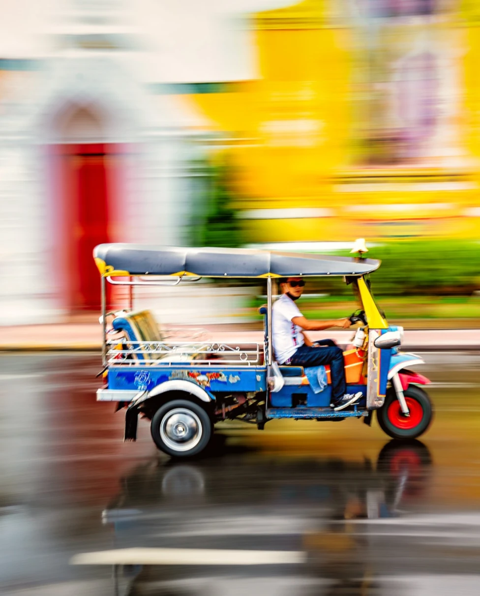 A man driving a tuk-tuk with an unfocused blurred colorful background of a street and yellow building