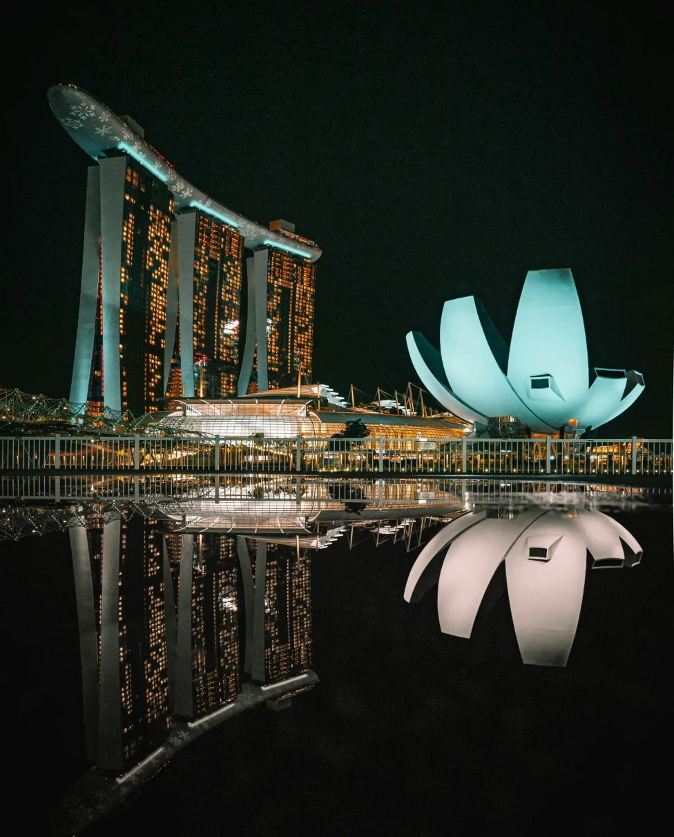 The image shows the Marina Bay Sands Hotel and ArtScience Museum in Singapore at night, with their reflections visible on the water’s surface.