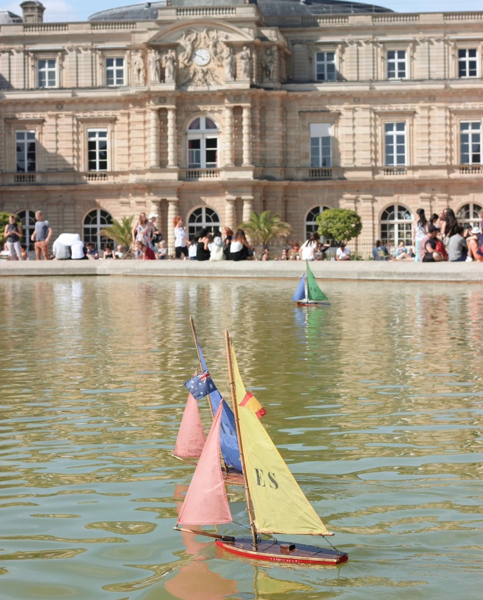 Vibrant model sailboats glide on water, with a grand edifice backdrop and onlookers at the water’s edge.