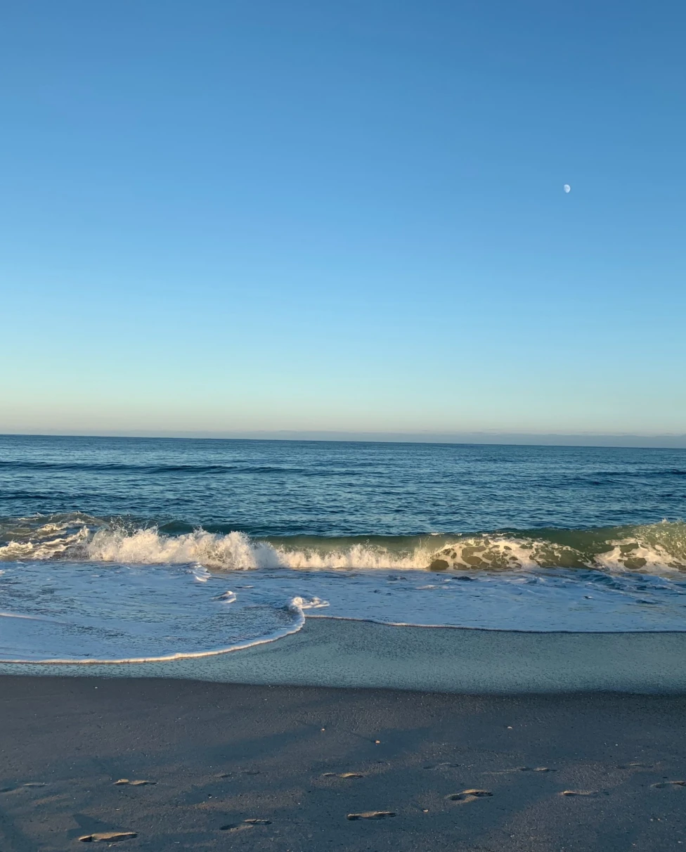 A wave breaks on a sandy beach with clear blue water in the foreground.