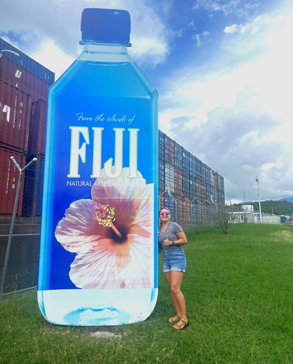 Danielle standing next to a large advertisement of a Fiji water bottle, with a clear sky and containers in the background.