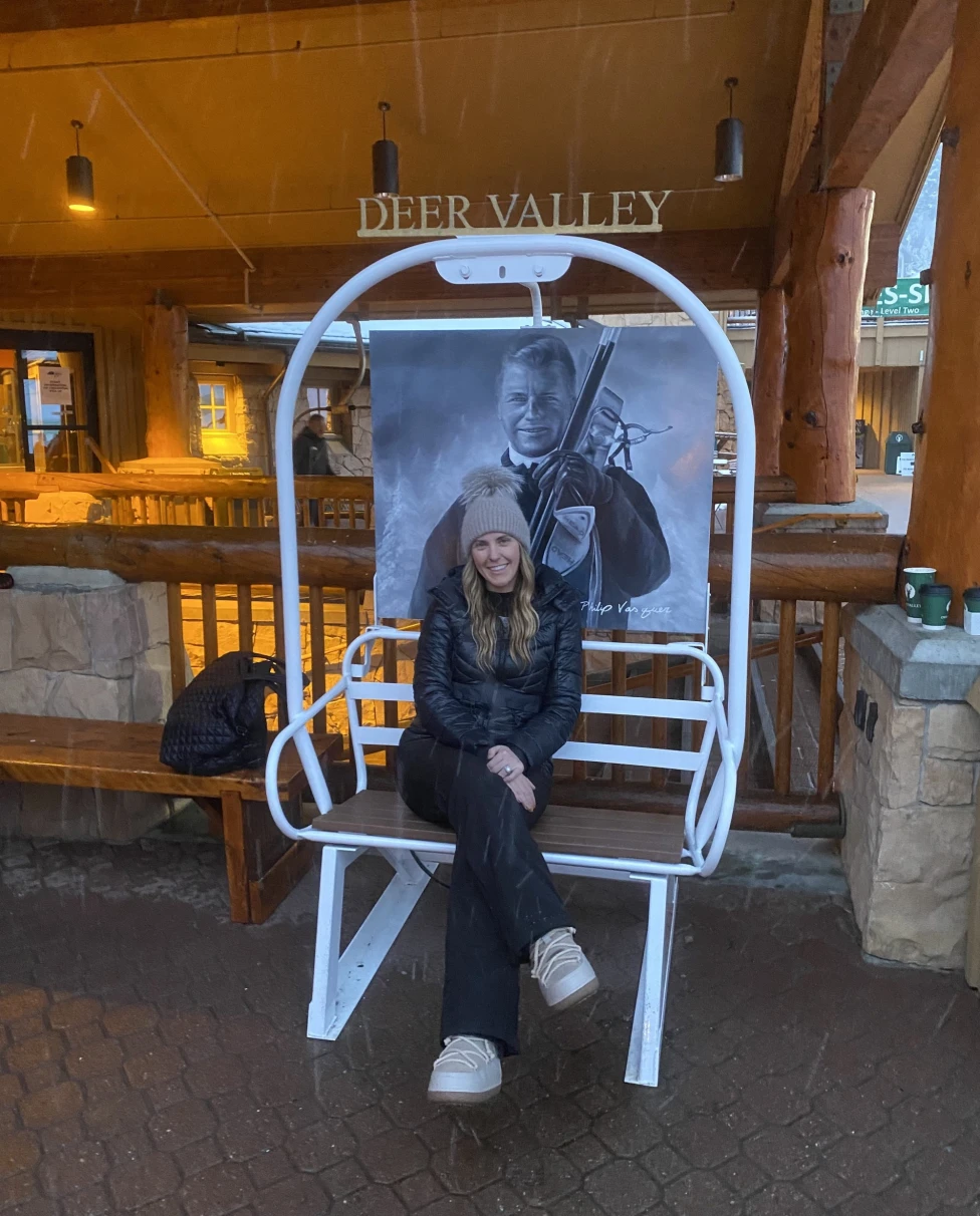 A woman sitting on a white ski chair in a lobby with a Deer Valley sign in the background.