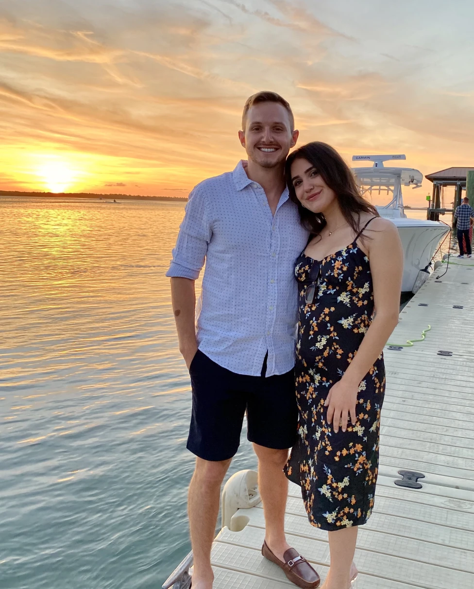A couple posing in front of a sunset over a body of water while standing on top of a dock.