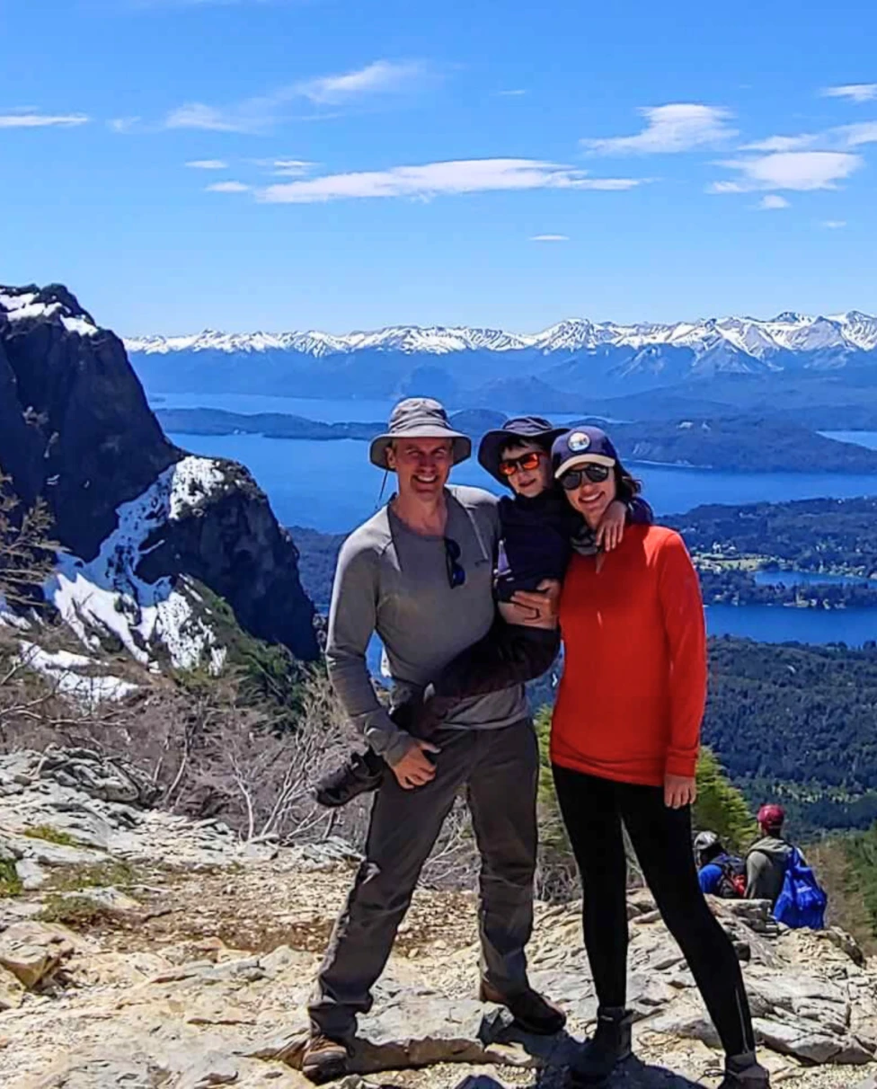 Two individuals are standing on a mountain trail with a panoramic view of a lake and mountains in the background.