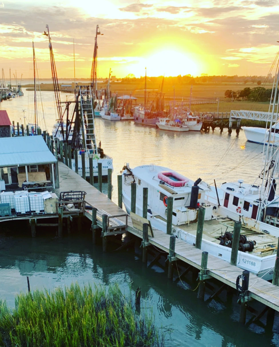 A Charleston sea port along the docks is dotted with boats as the sun sets behind the horizon at dusk.