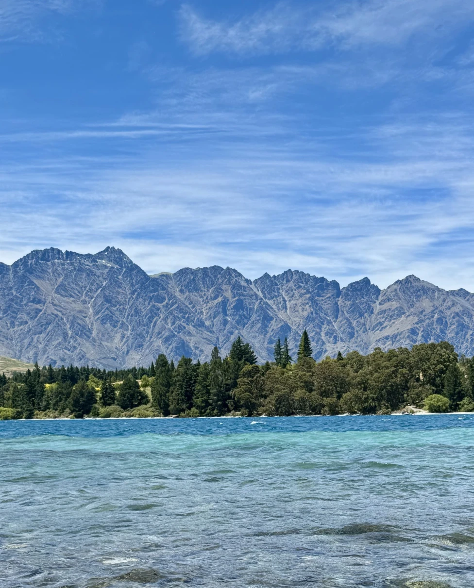 A vibrant landscape with a clear blue lake in the foreground and rugged mountains in the background under a partly cloudy sky.