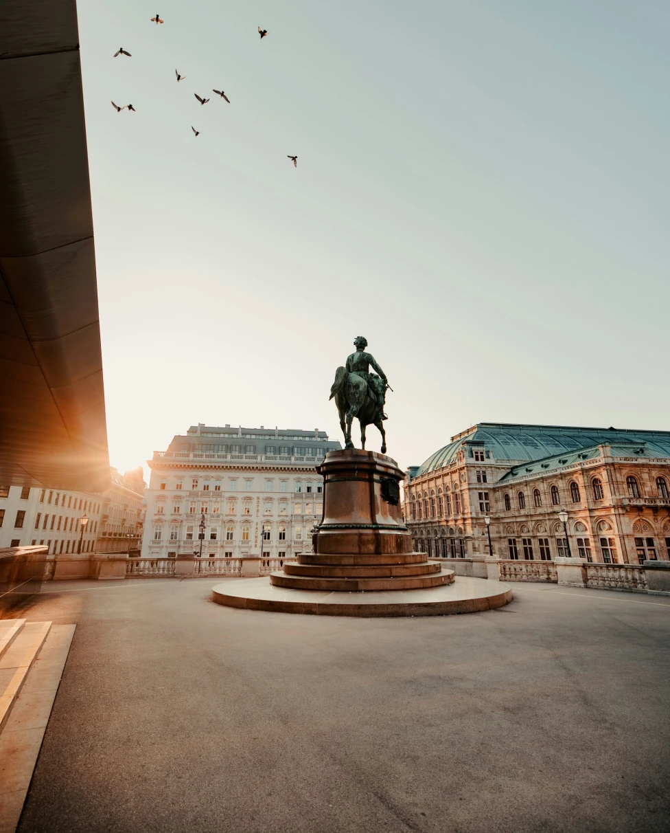 A statue of a mounted figure with a flock of birds flying above, set against grand buildings in warm sunlight.