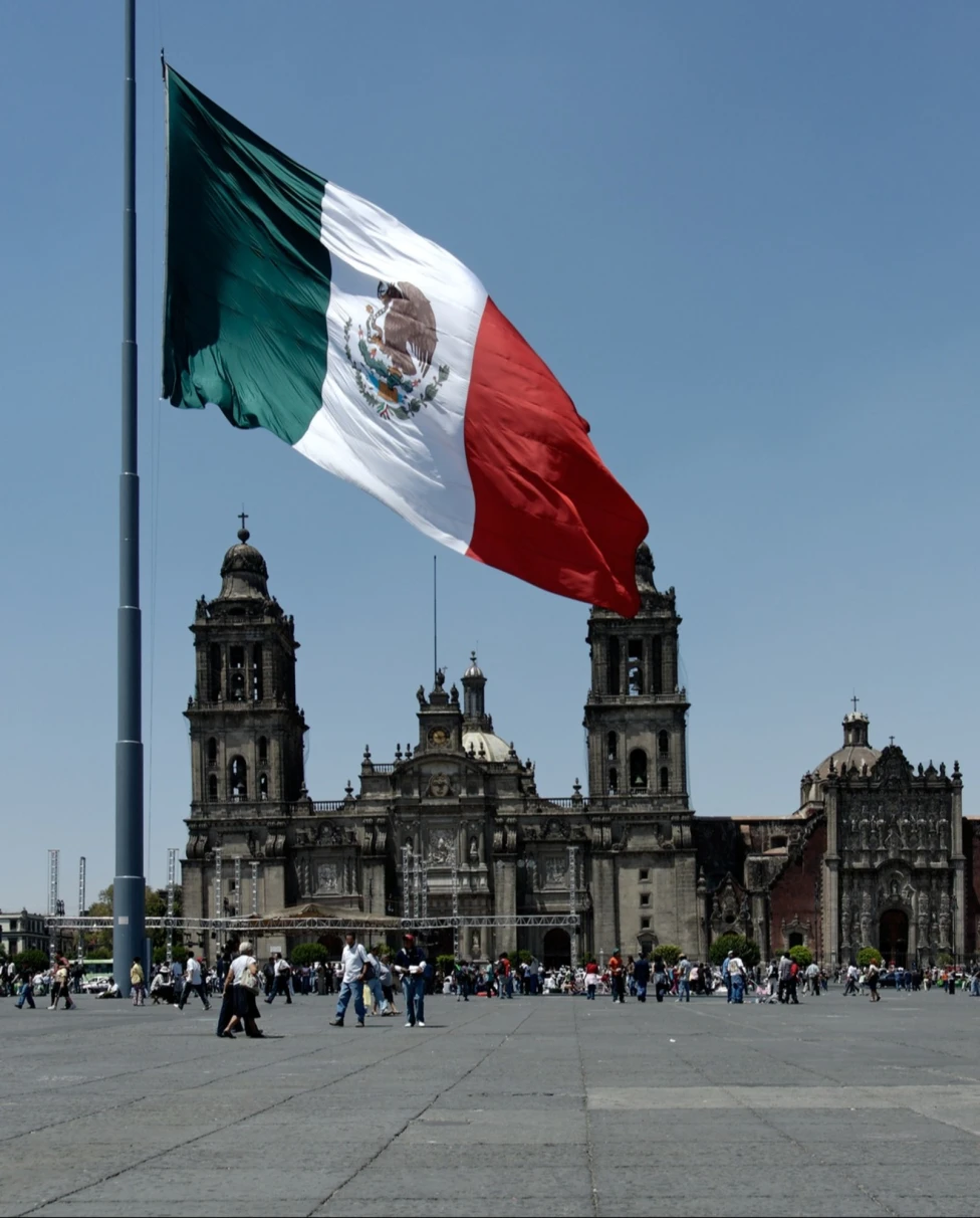 A view of the flag in Mexico in front of people walking near a grand stone building.
