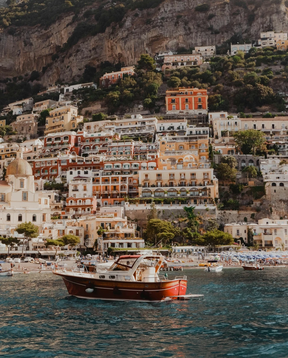 A view of a red and white boat sailing on the water in front of a coastal Italian town.