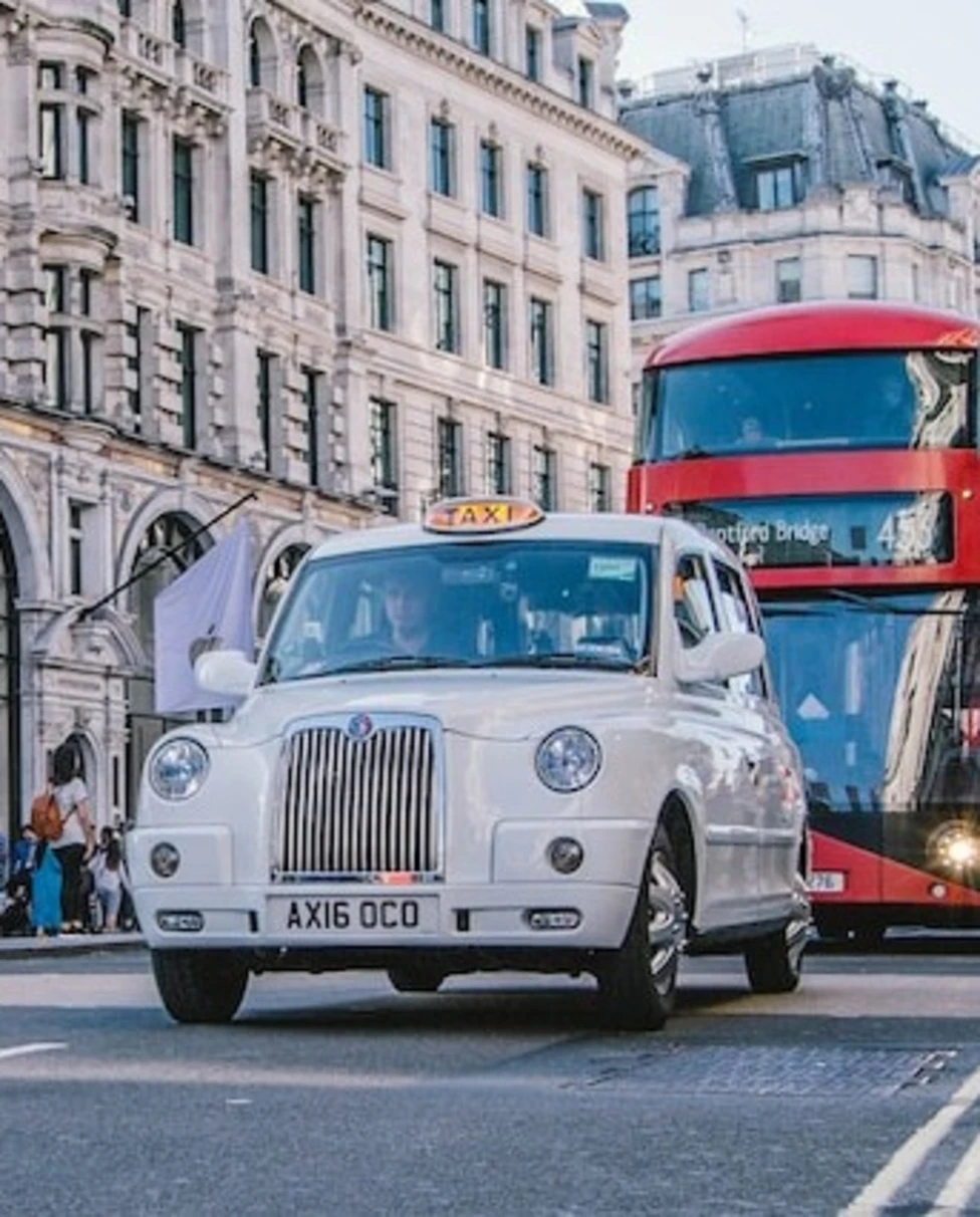 A red colored double decker and a white car on the road in London.