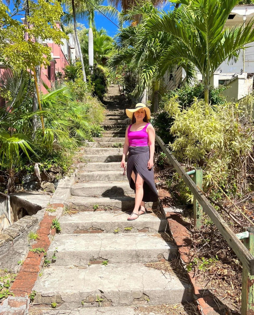 A person in a wide-brimmed sun hat and bright pink top stands on a sunlit stone staircase surrounded by lush tropical greenery.