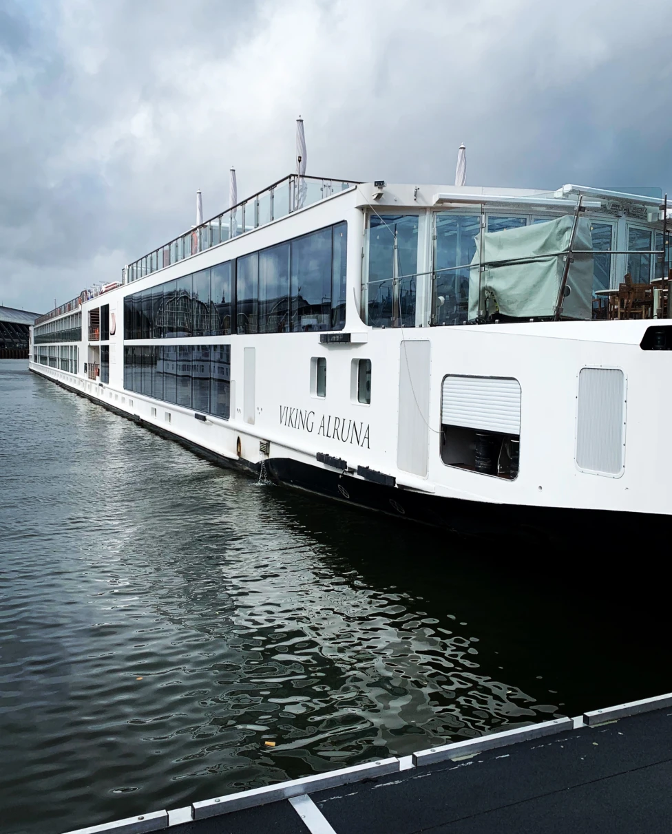 A large white riverboat named ‘Viking Alruna’ is docked on a calm body of water with a bridge in the background under an overcast sky.