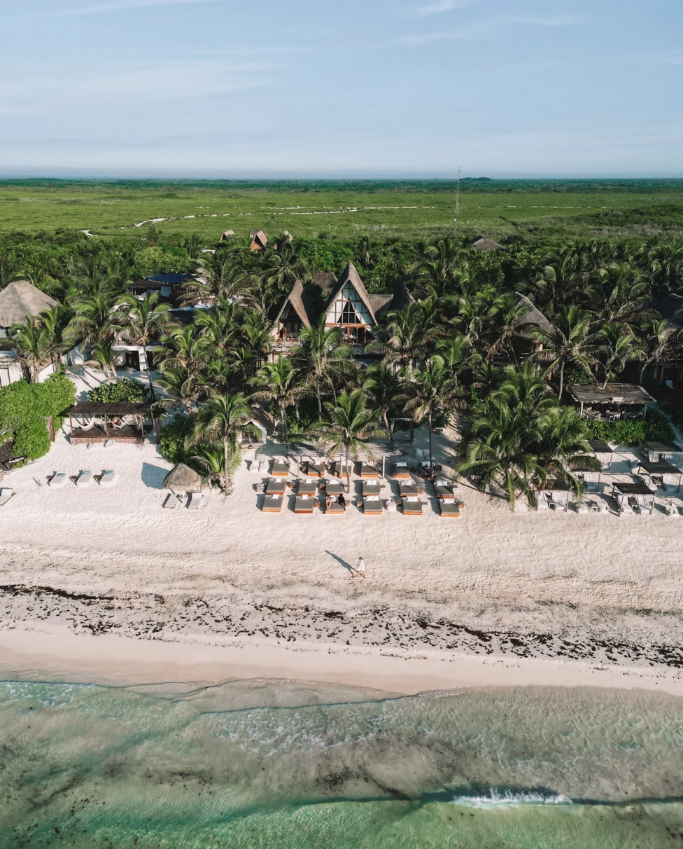 This image depicts an aerial view of a beach, ocean and villas aligned in row on the sandy shore. There is one large a-frame building in the background surrounded by lush green trees and foliage.